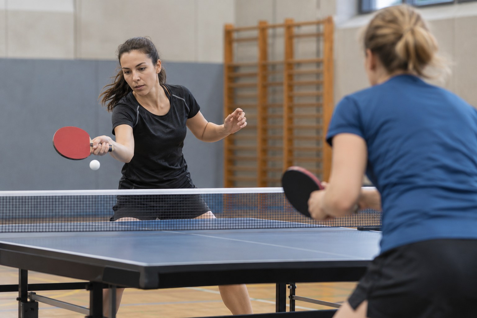 Zwei Spielerinnen trainieren an einer Tischtennisplatte in einer Sporthalle.