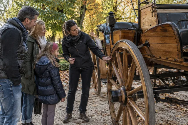 Eine Familie betrachtet mit einem Guide eine historische Kutsche im Grünen.